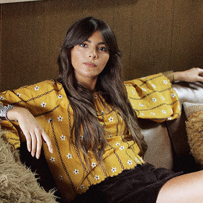 Hero banner for The Hippie Shake's boho and 70s blouses and tops - Model wears yellow embroidered smock top sat on sofa.