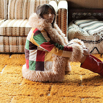 Hero banner for boho and 70s style coats and jackets from The Hippie Shake - Model wearing patchwork Penny Lane coat sat on yellow rug.