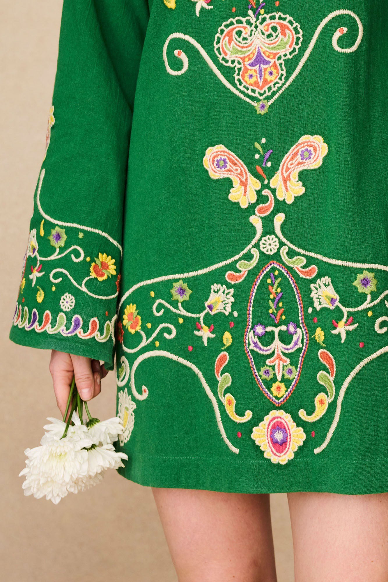 Close-up of the hem of The Hippie Shake's green embroidered mini dress with intricate floral stitching and model holding white flowers.