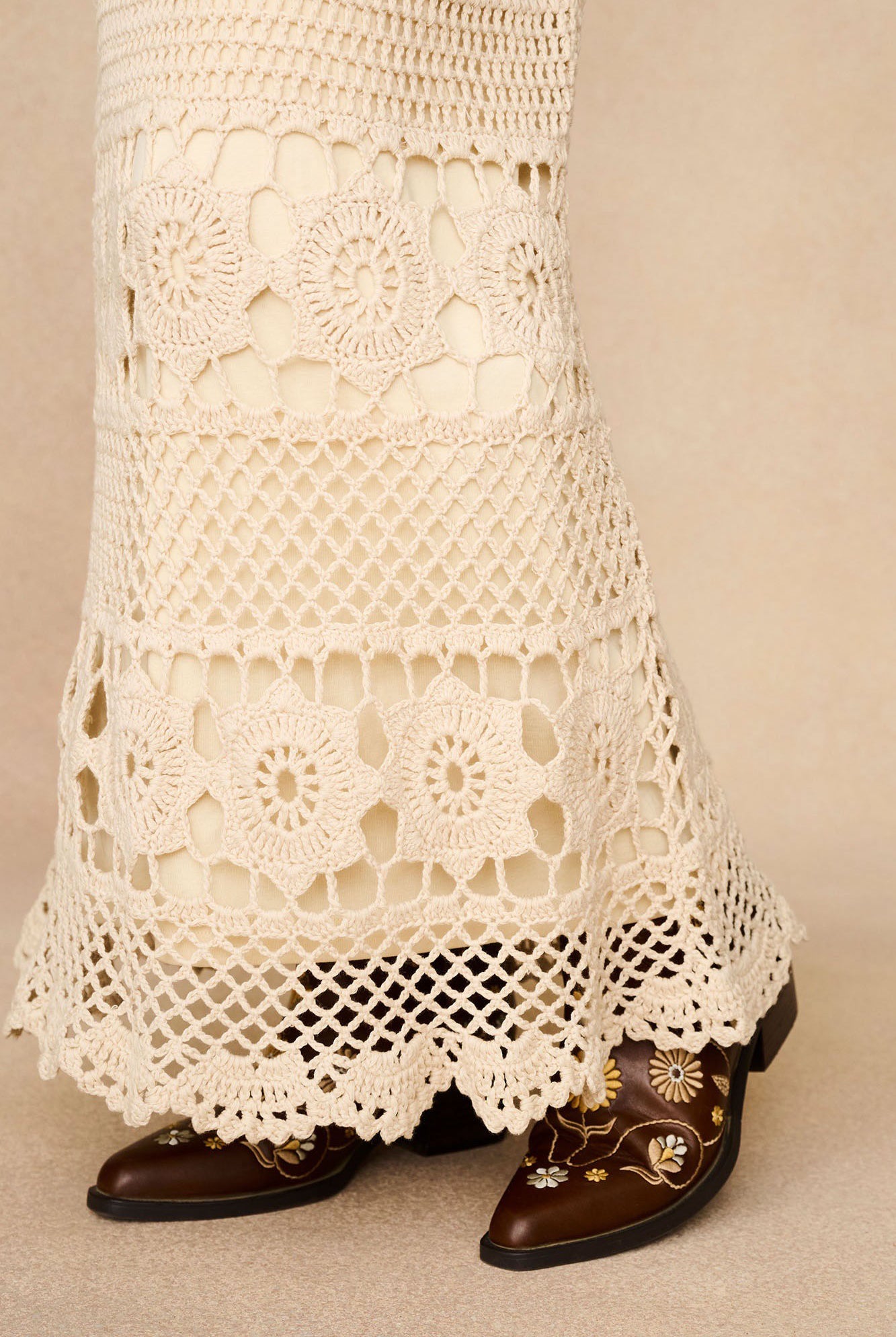 Detail shot of The Hippie Shake's vintage style cream maxi skirt's floral crochet hem worn with embroidered brown boots.