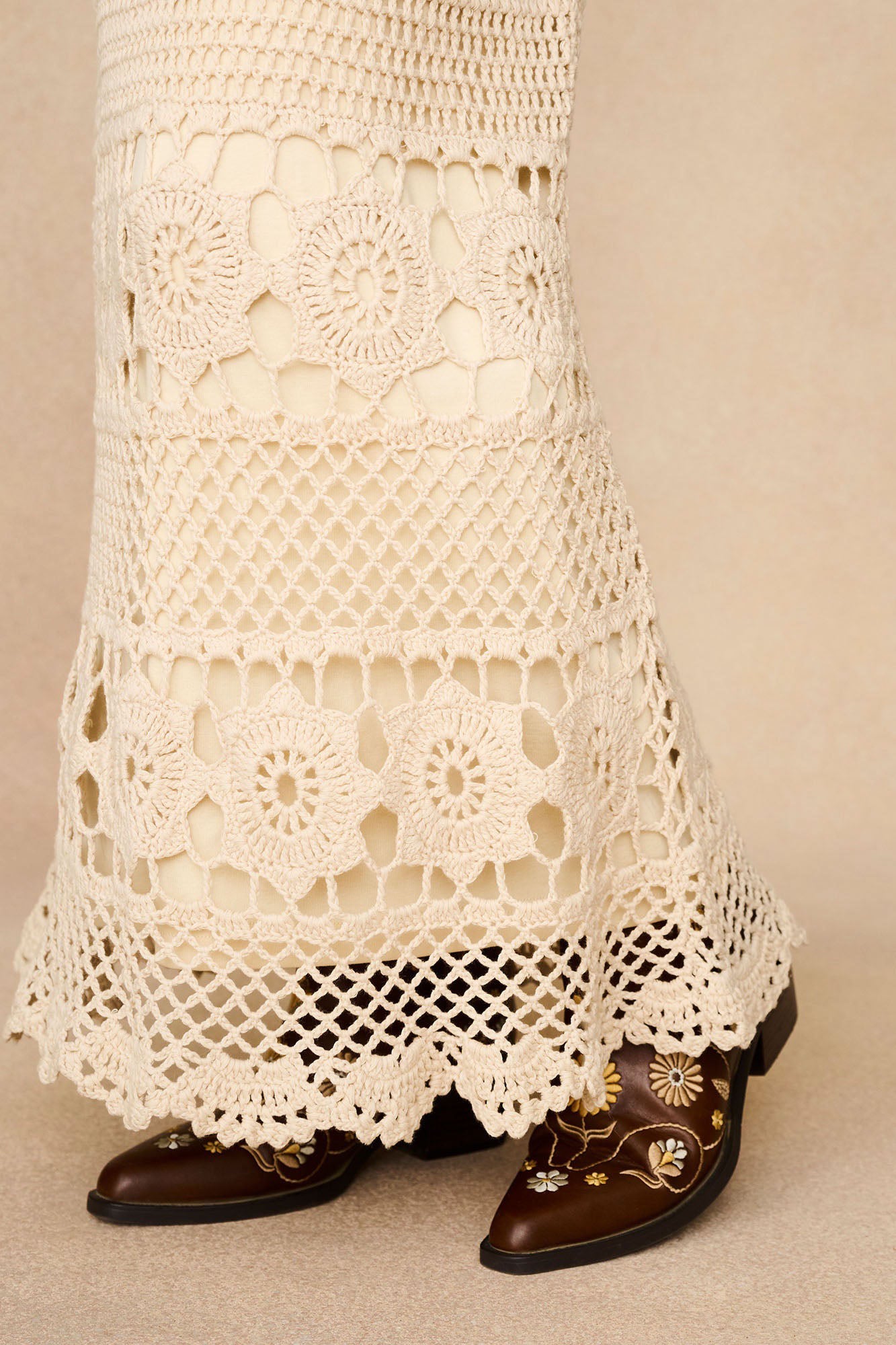 Detail shot of The Hippie Shake's vintage style cream maxi skirt's floral crochet hem worn with embroidered brown boots.