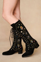 Model wearing The Hippie Shake's black embroidered knee-high boots with floral detailing, legs facing left