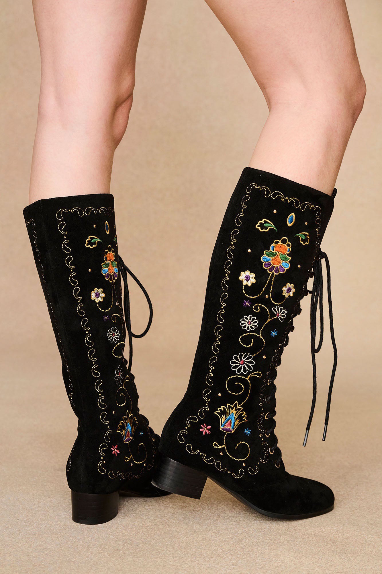 Model styled in The Hippie Shake's black embroidered boots with lace-up fronts and retro floral embroidery, legs angled right