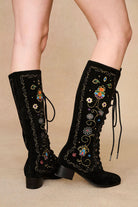 Model styled in The Hippie Shake's black embroidered boots with lace-up fronts and retro floral embroidery, legs angled right