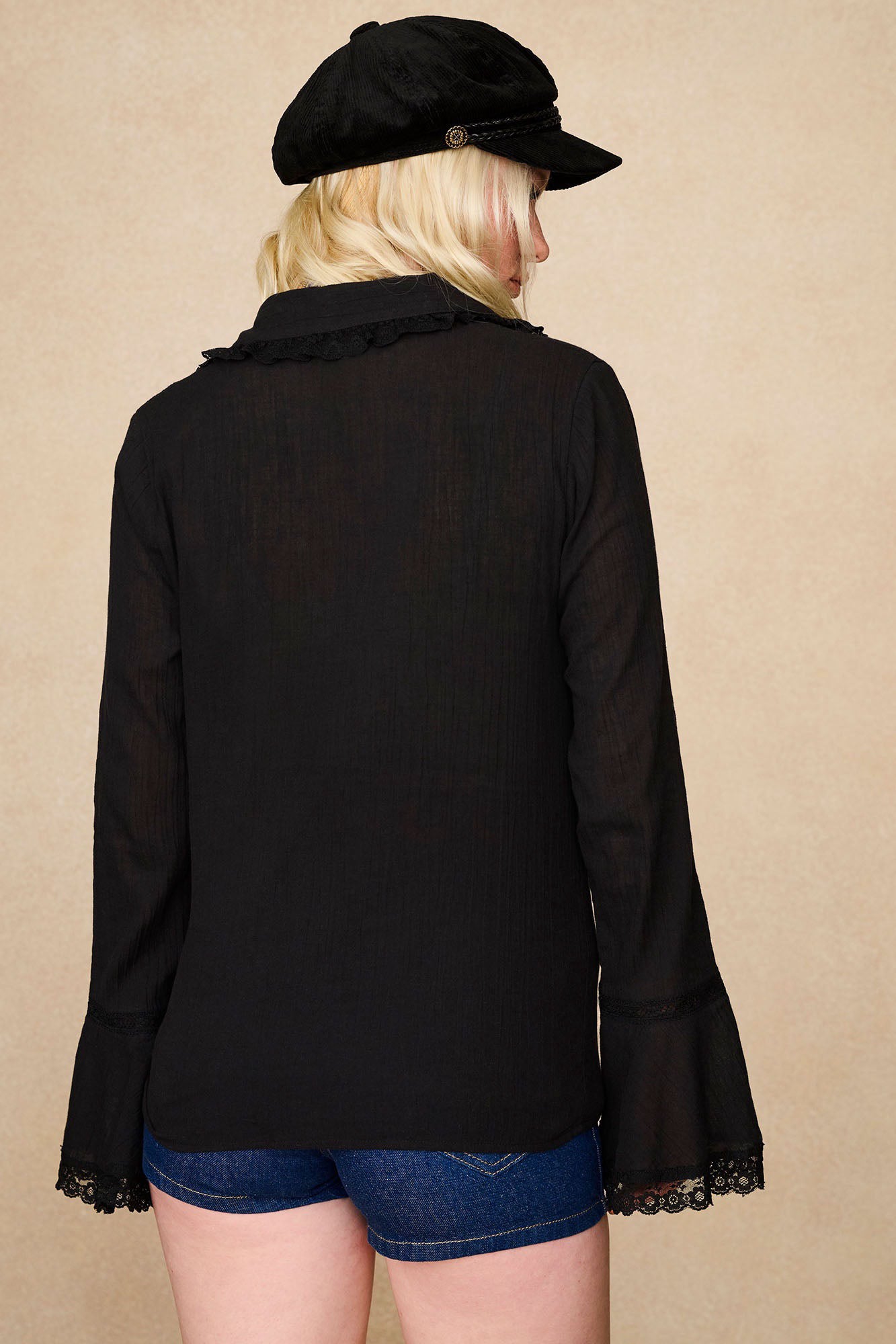 Back view of model in The Hippie Shake's black lace trim blouse with lace trim cuffs and collar, styled with denim shorts and black baker hat.