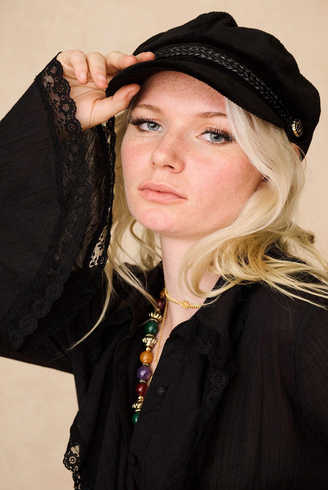 Close-up of model in The Hippie Shake's black ruffled blouse, lifting our black baker hat, styled with layered beaded necklaces.