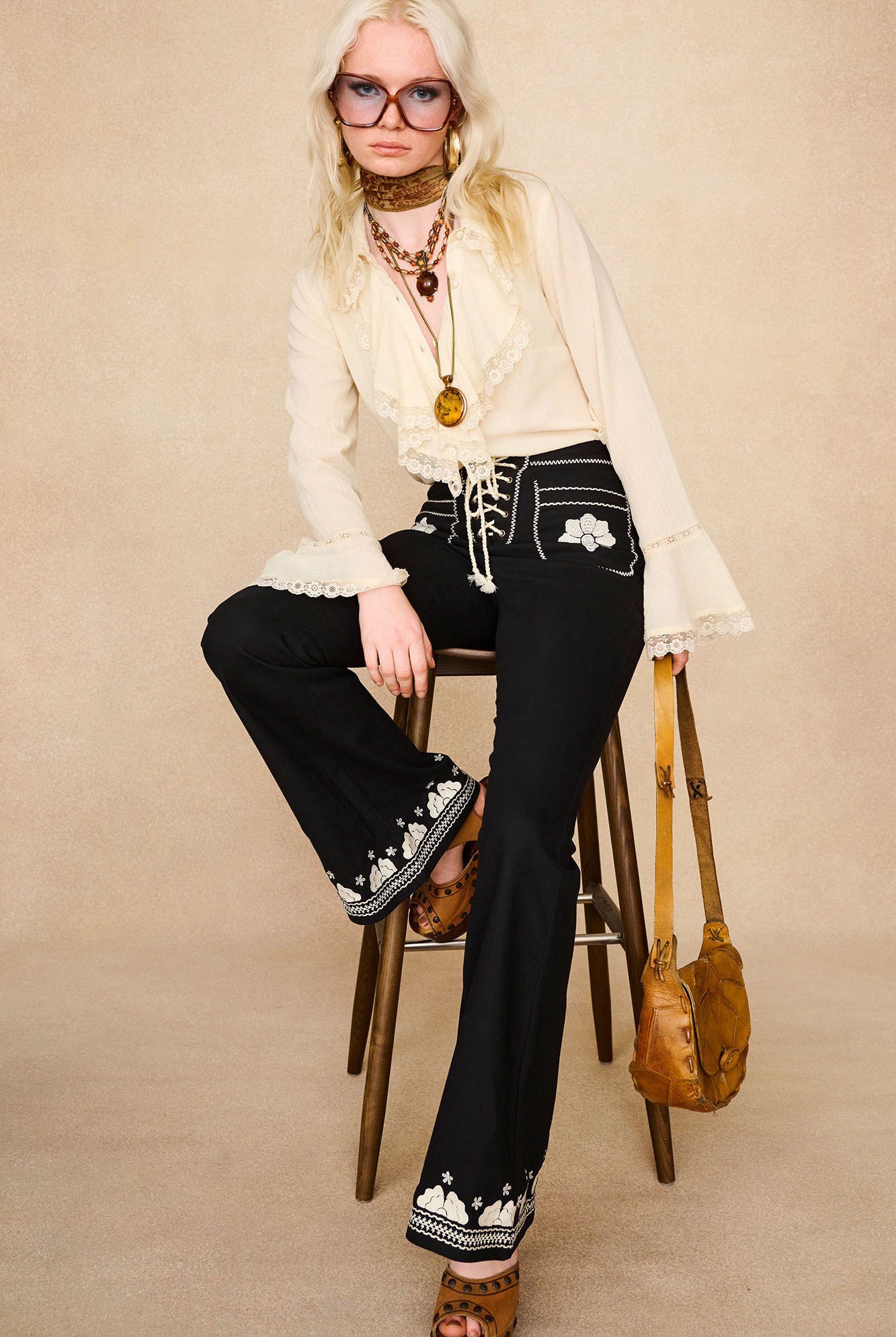 Full-length shot of model sitting wearing The Hippie Shake's black embroidered floral flared trousers with cream lace trim ruffle blouse.
