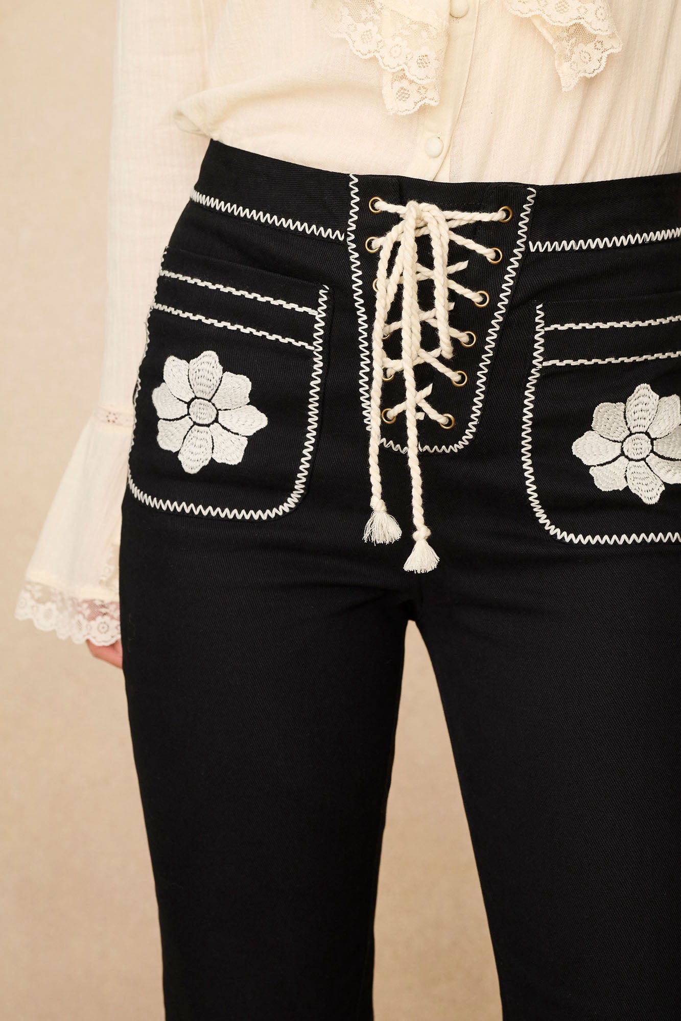 Close-up of The Hippie Shake's 70s style black floral flared trousers featuring the intricate embroidery and lace up detail at the waist.