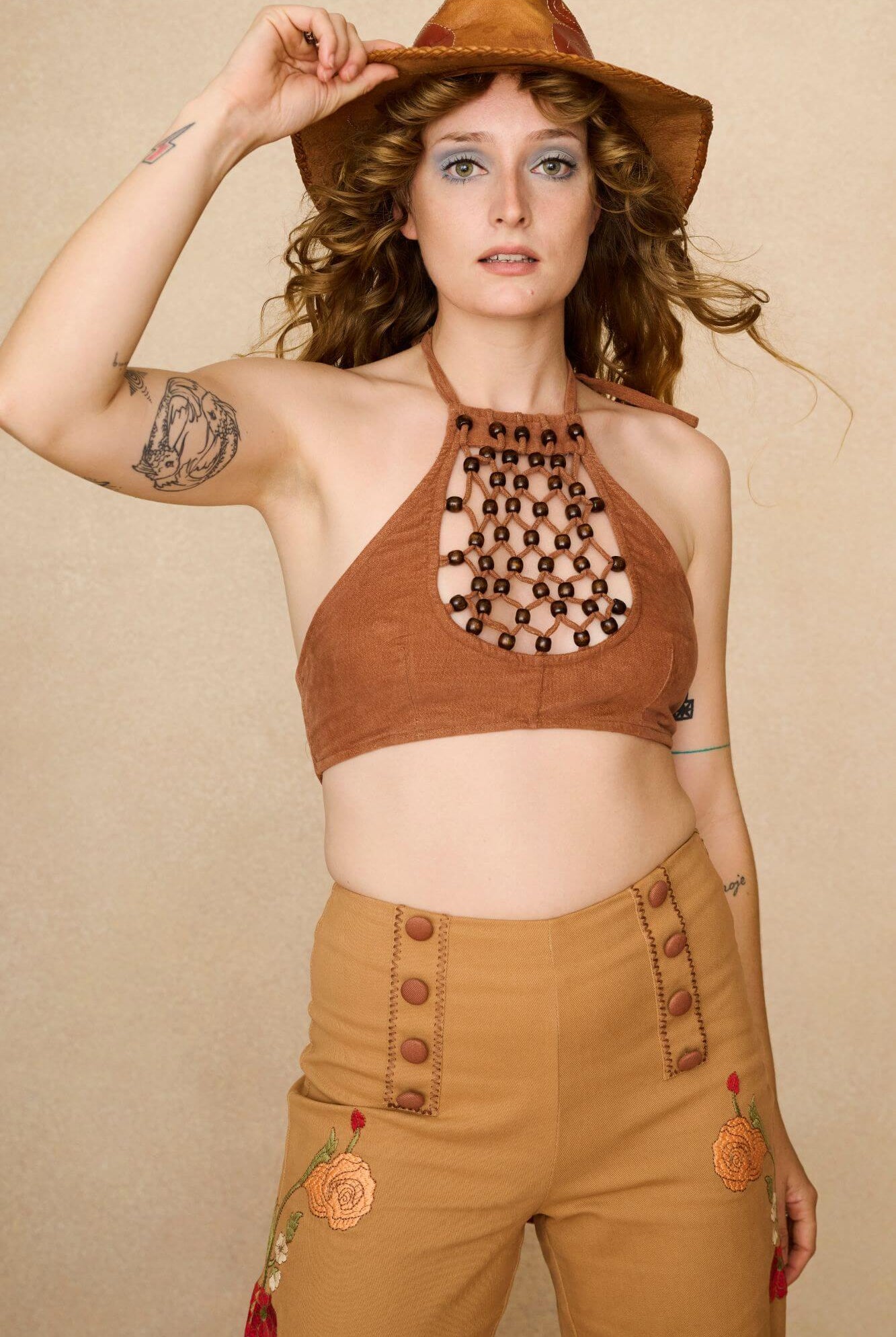 Woman wearing a 70s style brown halter top with embellishments and high-waisted pants against a beige background - front view