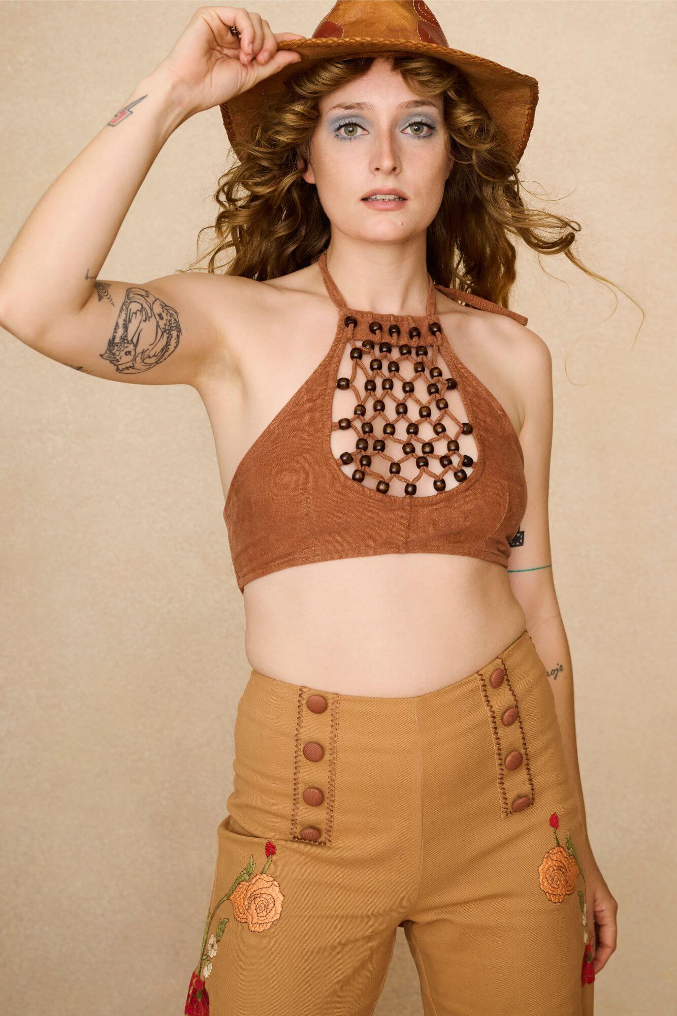 Woman wearing a 70s style brown halter top with embellishments and high-waisted pants against a beige background - front view