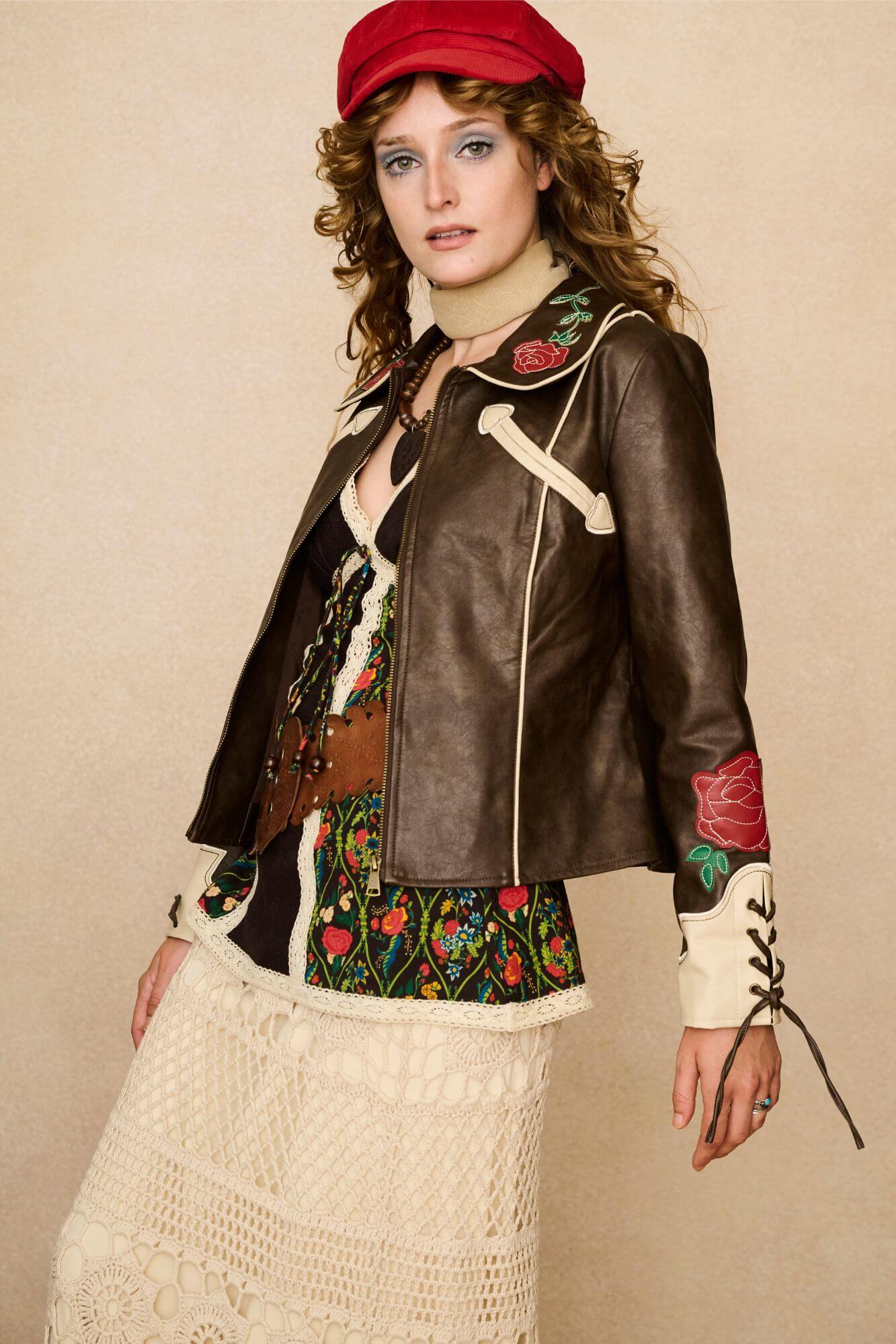 Model wearing a brown 70s style faux leather jacket with red floral embroidery against a beige background - outfit with movement