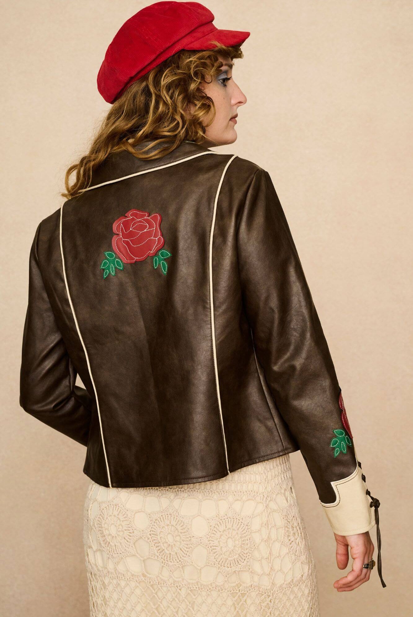 Model wearing a brown 70s style faux leather jacket with red floral embroidery against a beige background - back view