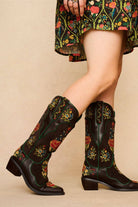 Brown Western style embroidered boots with floral patterns worn by a model on a beige background - side view
