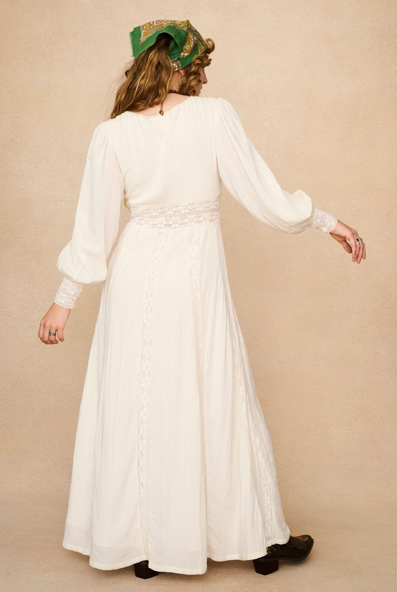 Woman in a long cream lace floaty 70s style maxi dress against a beige background - back view