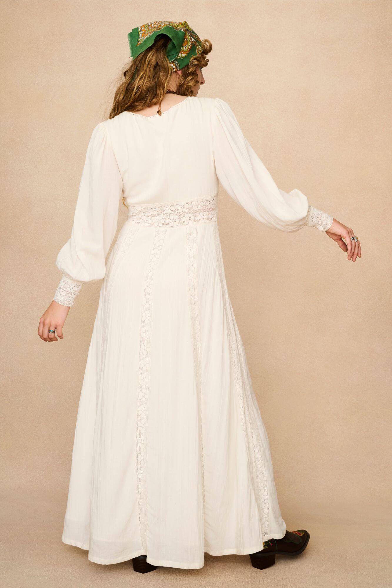 Woman in a long cream lace floaty 70s style maxi dress against a beige background - back view