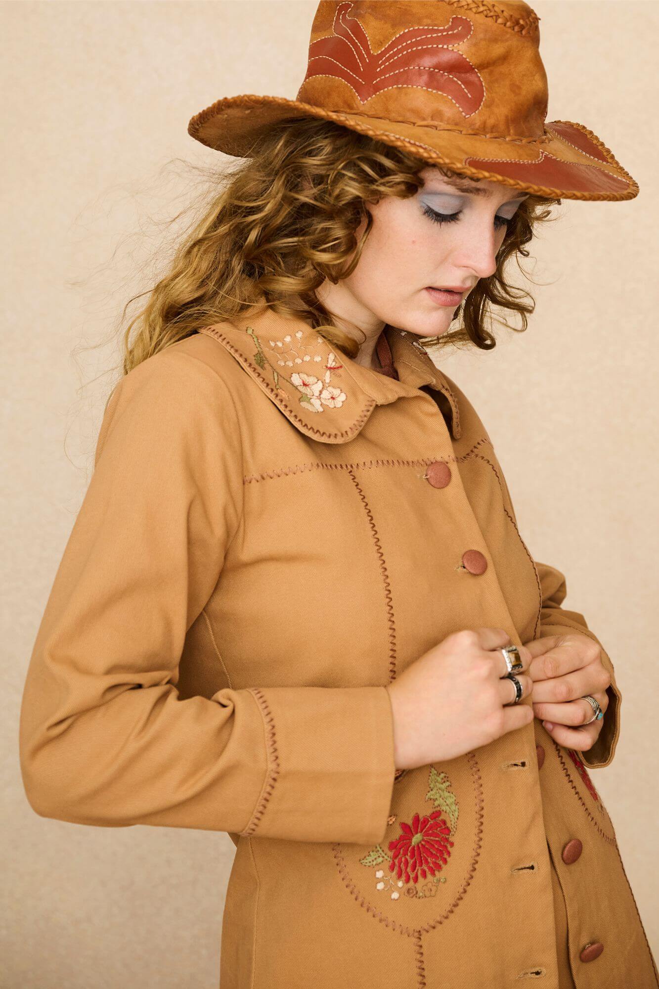 Model wearing a tan 70s style coat with embroidered floral patches against a beige background - close up fit