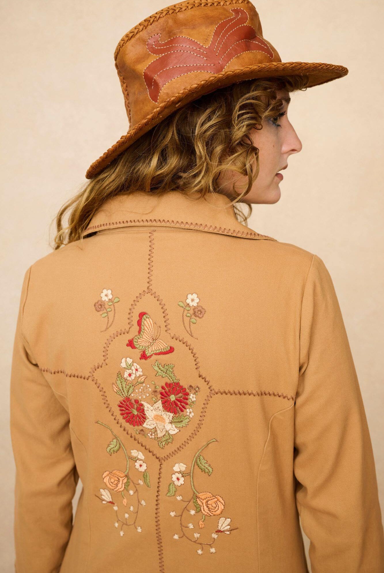 Model wearing a tan 70s style coat with embroidered floral patches against a beige background - close up of back embroidery