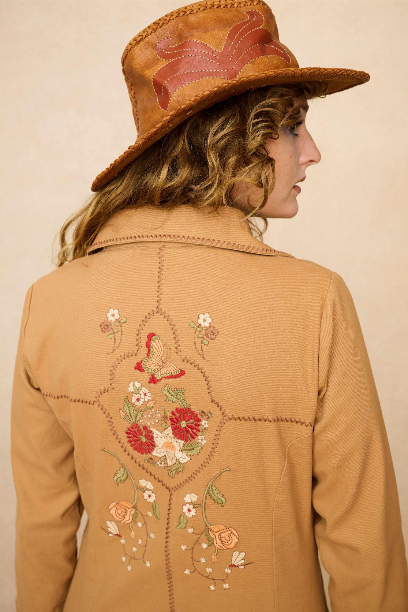 Model wearing a tan 70s style coat with embroidered floral patches against a beige background - close up of back embroidery