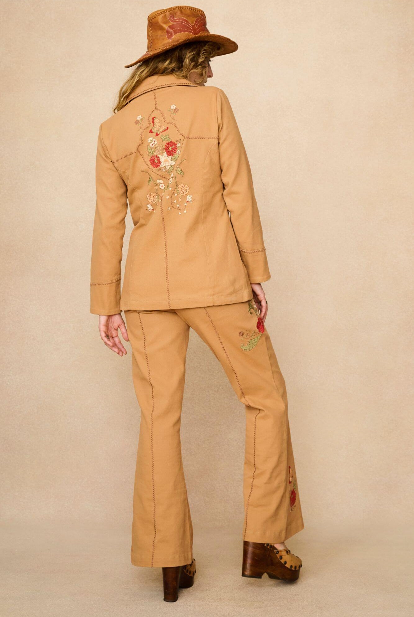 Model wearing a tan 70s style coat with embroidered floral patches against a beige background - back view