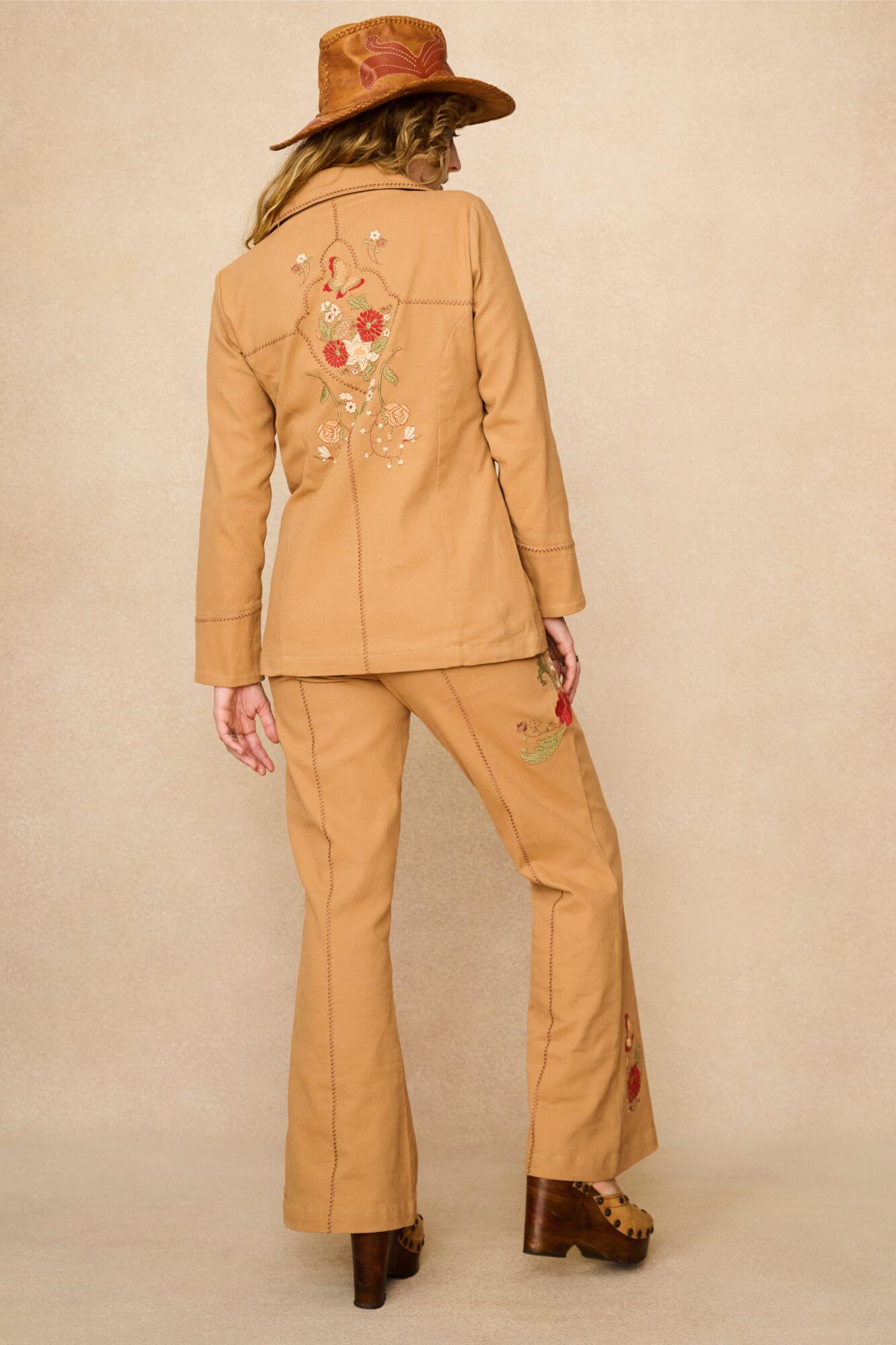 Model wearing a tan 70s style coat with embroidered floral patches against a beige background - back view
