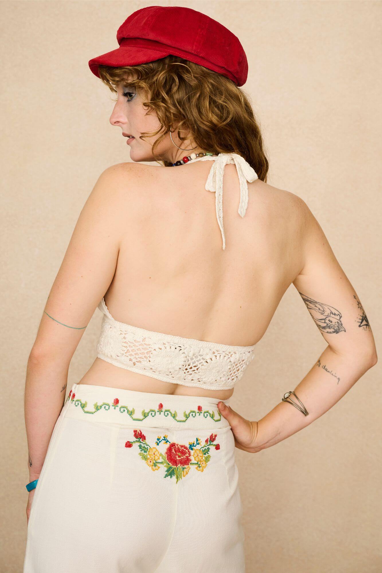 Woman wearing a red cap, floral embroidered top, and colourful necklace against a beige background - back view