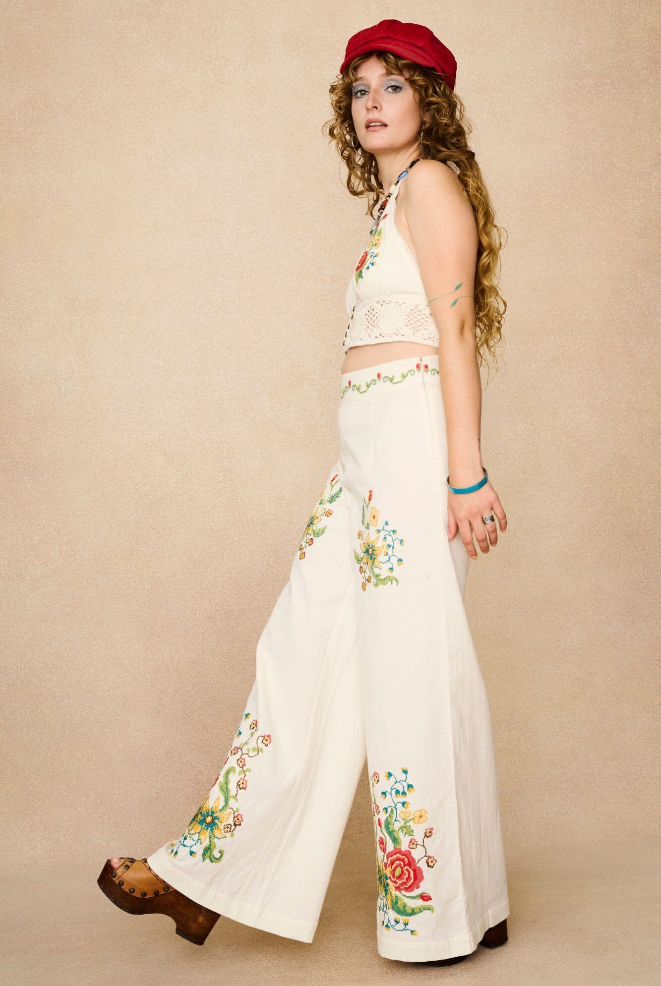 Model wearing white flared trousers with colourful floral embroidery on a beige background - side view