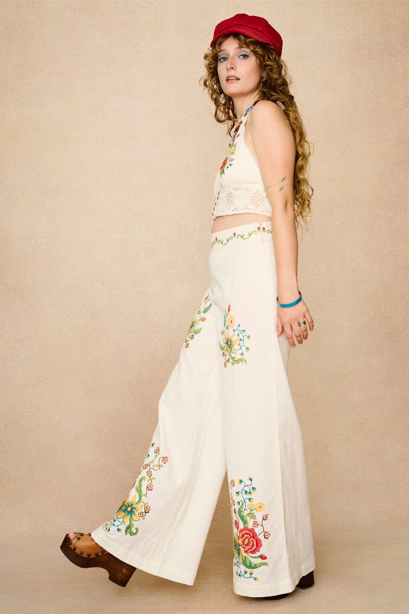 Model wearing white flared trousers with colourful floral embroidery on a beige background - side view