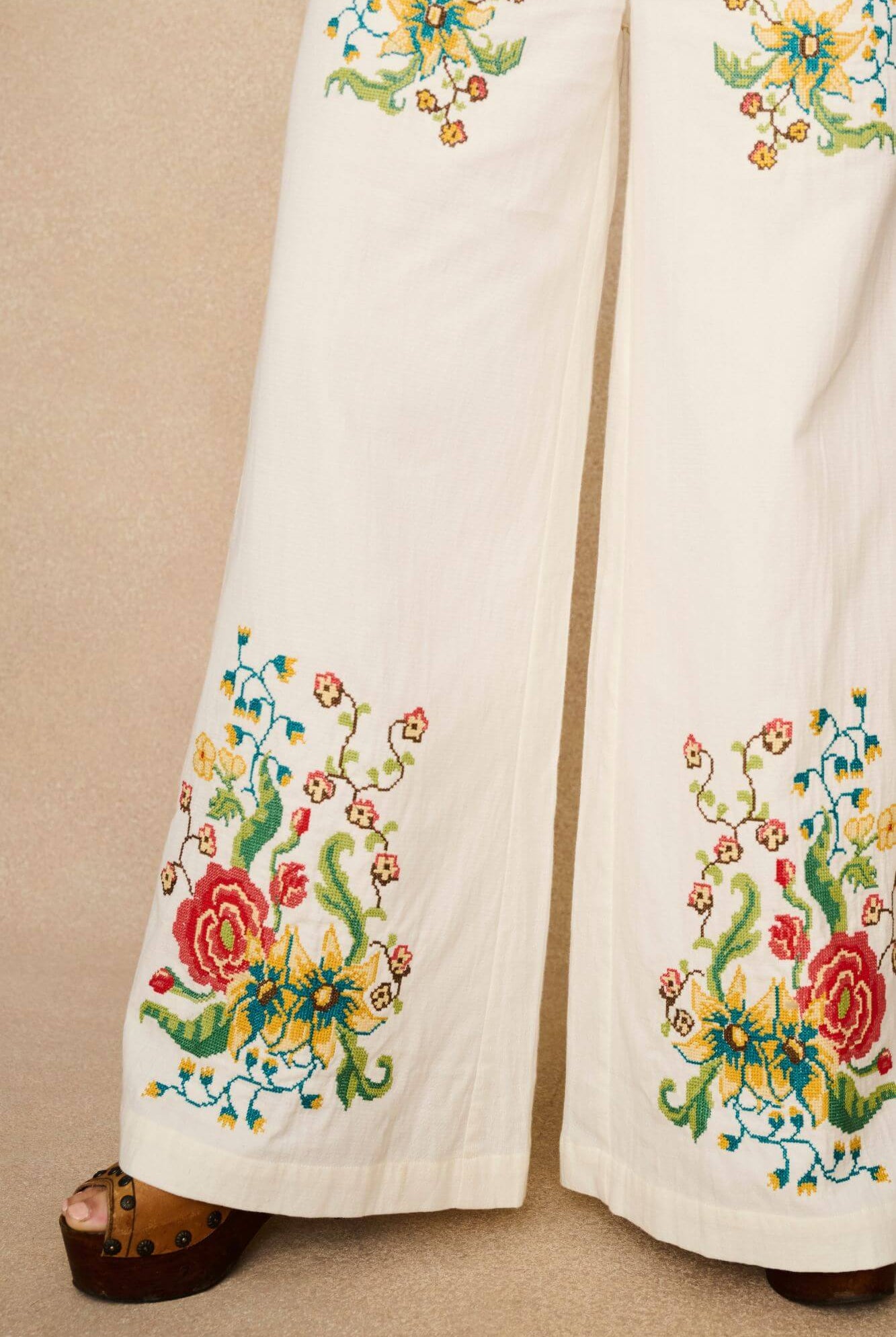 Model wearing white flared trousers with colourful floral embroidery on a beige background - close up of embroidery