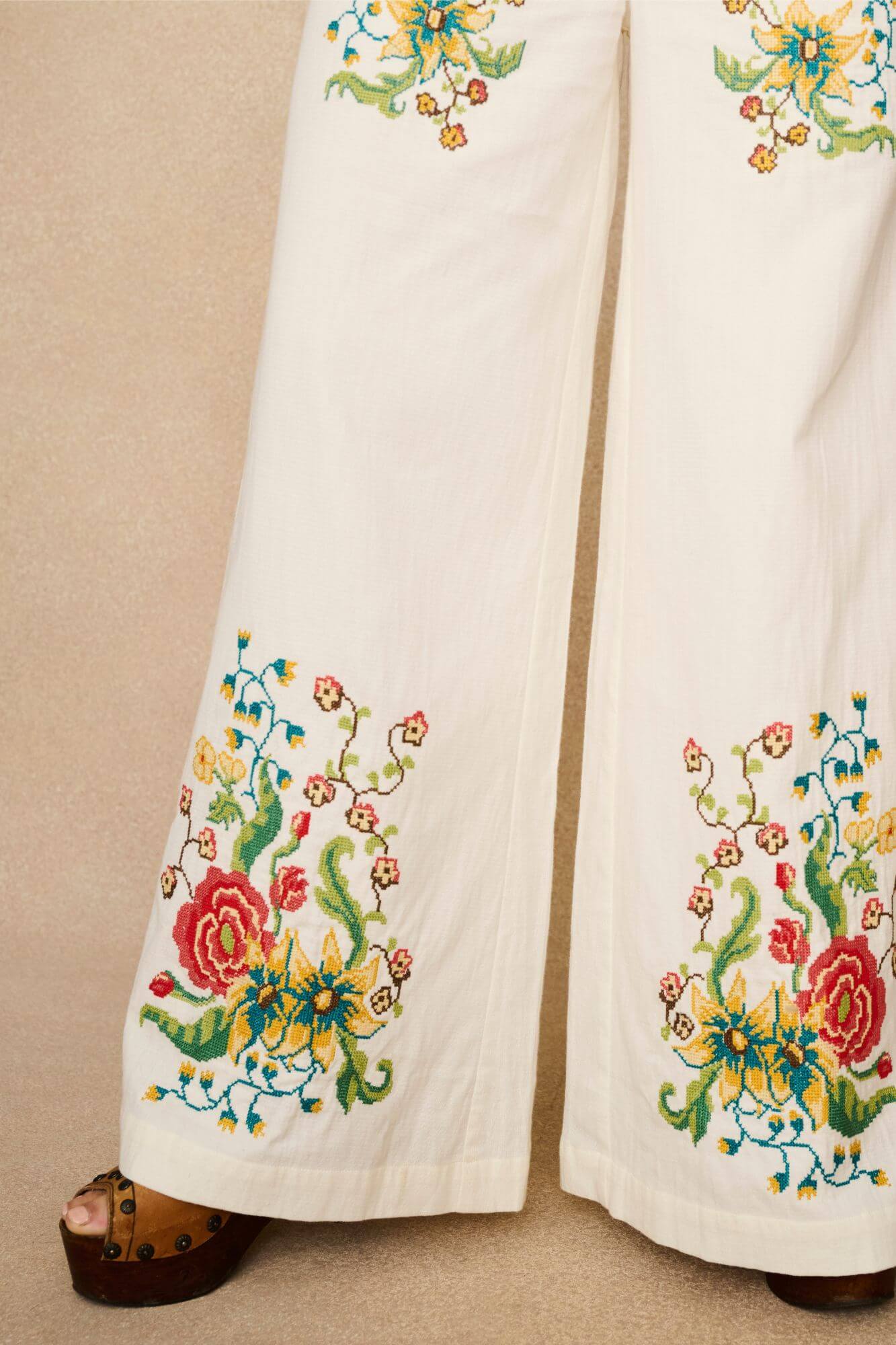 Model wearing white flared trousers with colourful floral embroidery on a beige background - close up of embroidery