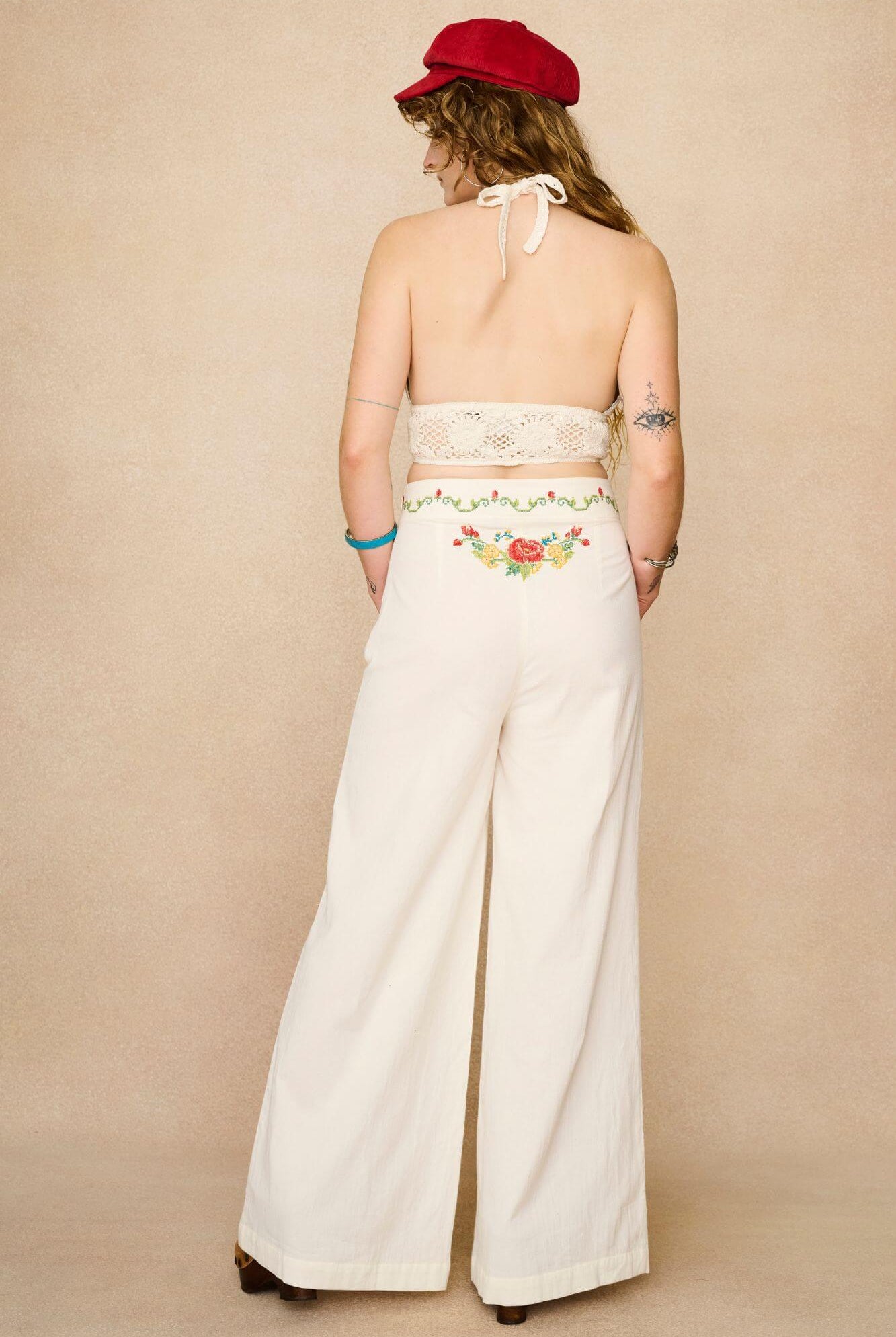 Model wearing white flared trousers with colourful floral embroidery on a beige background - back view