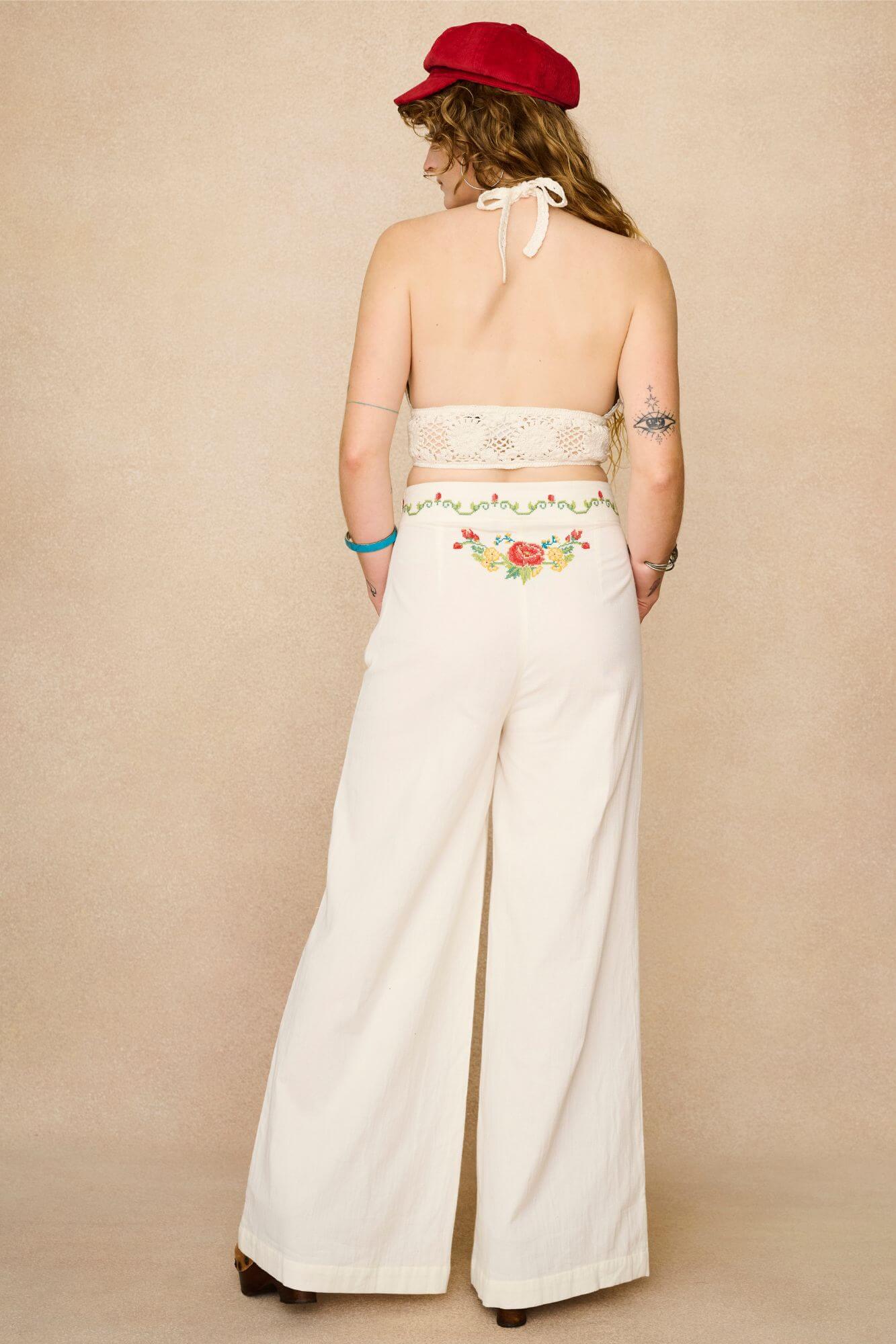 Model wearing white flared trousers with colourful floral embroidery on a beige background - back view