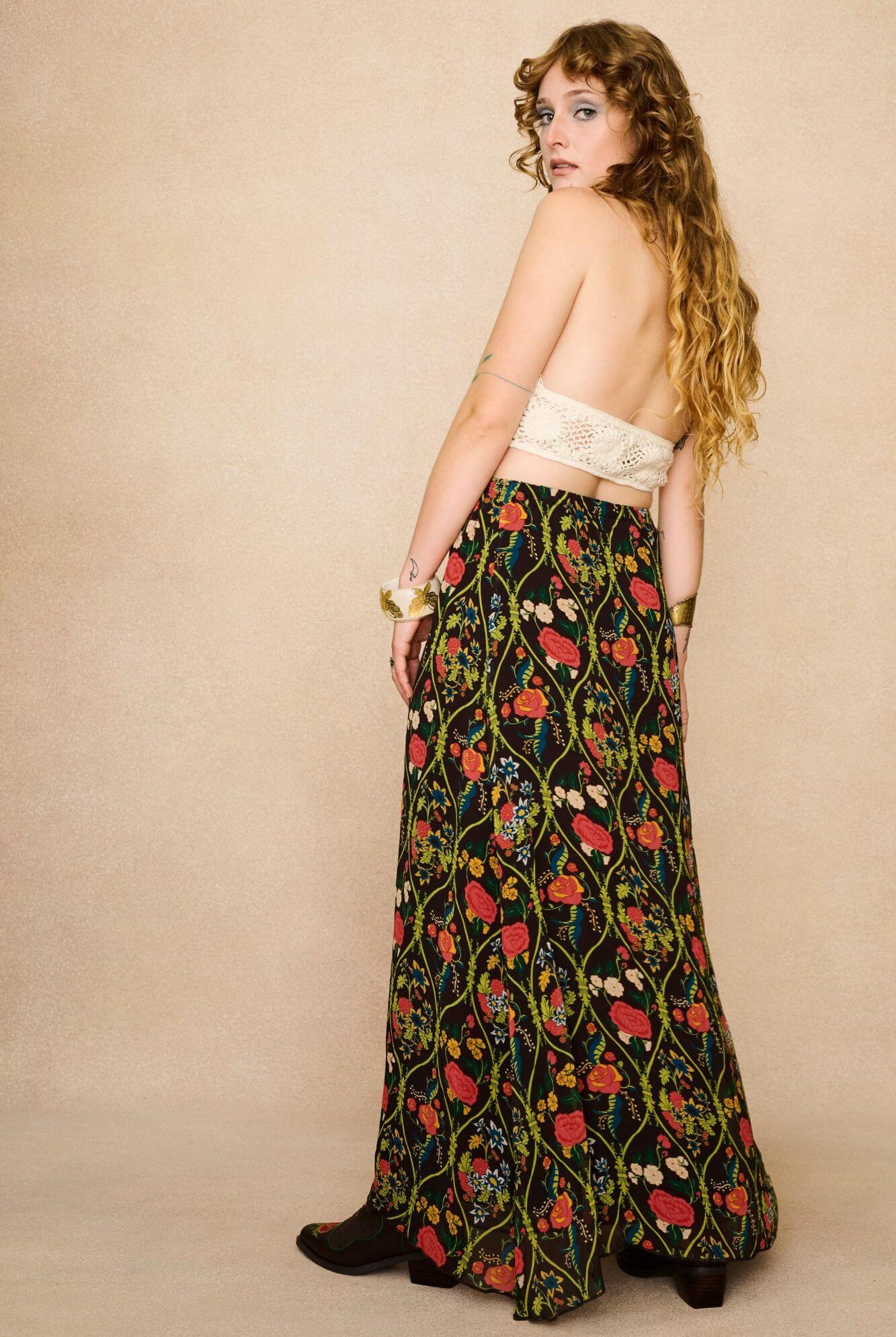 Model wearing a long 70s style floral skirt and white top with a beige background - back view