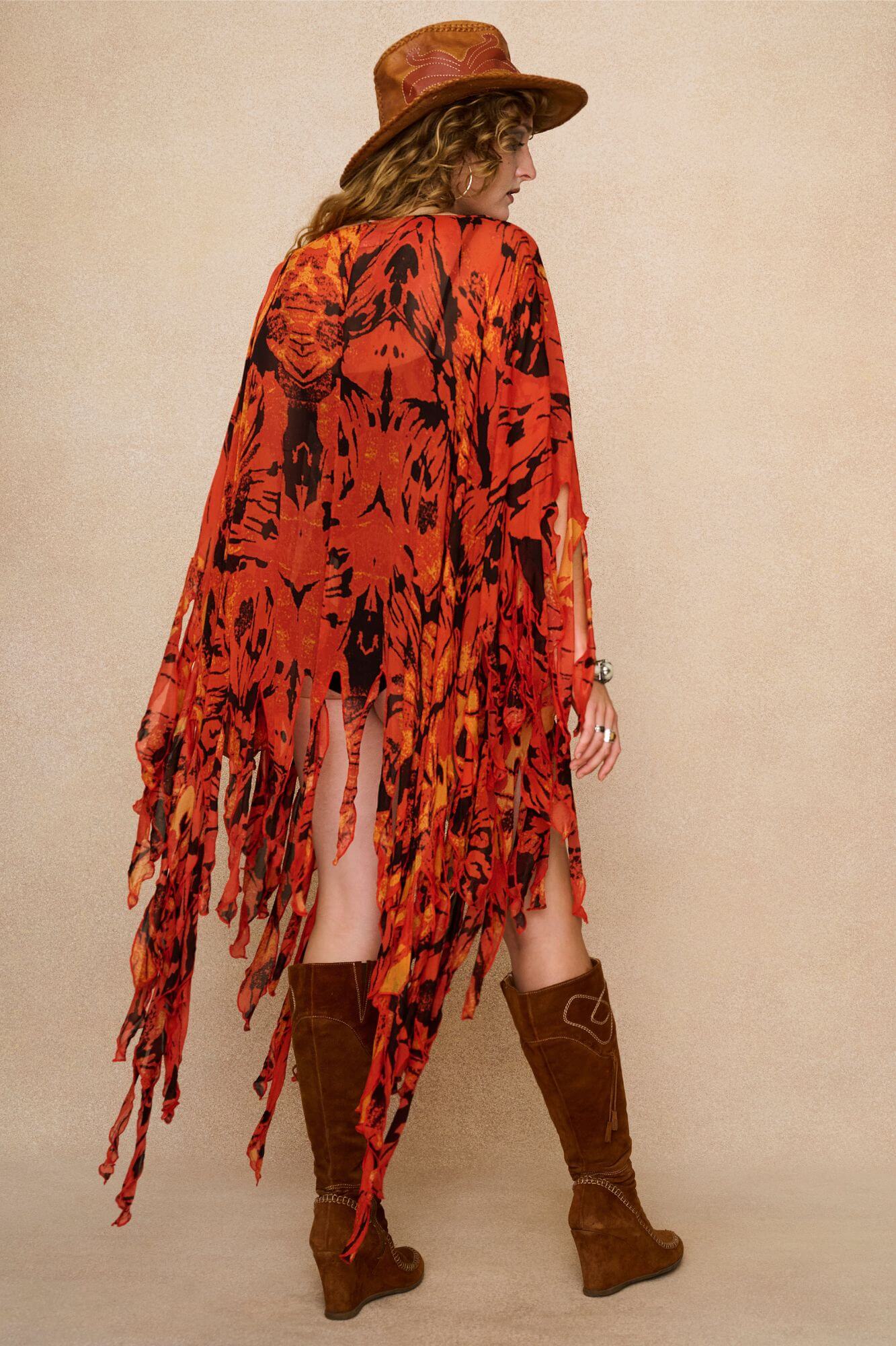 Model wearing a vibrant red and black patterned dress cape with fringe against a beige background - Back view