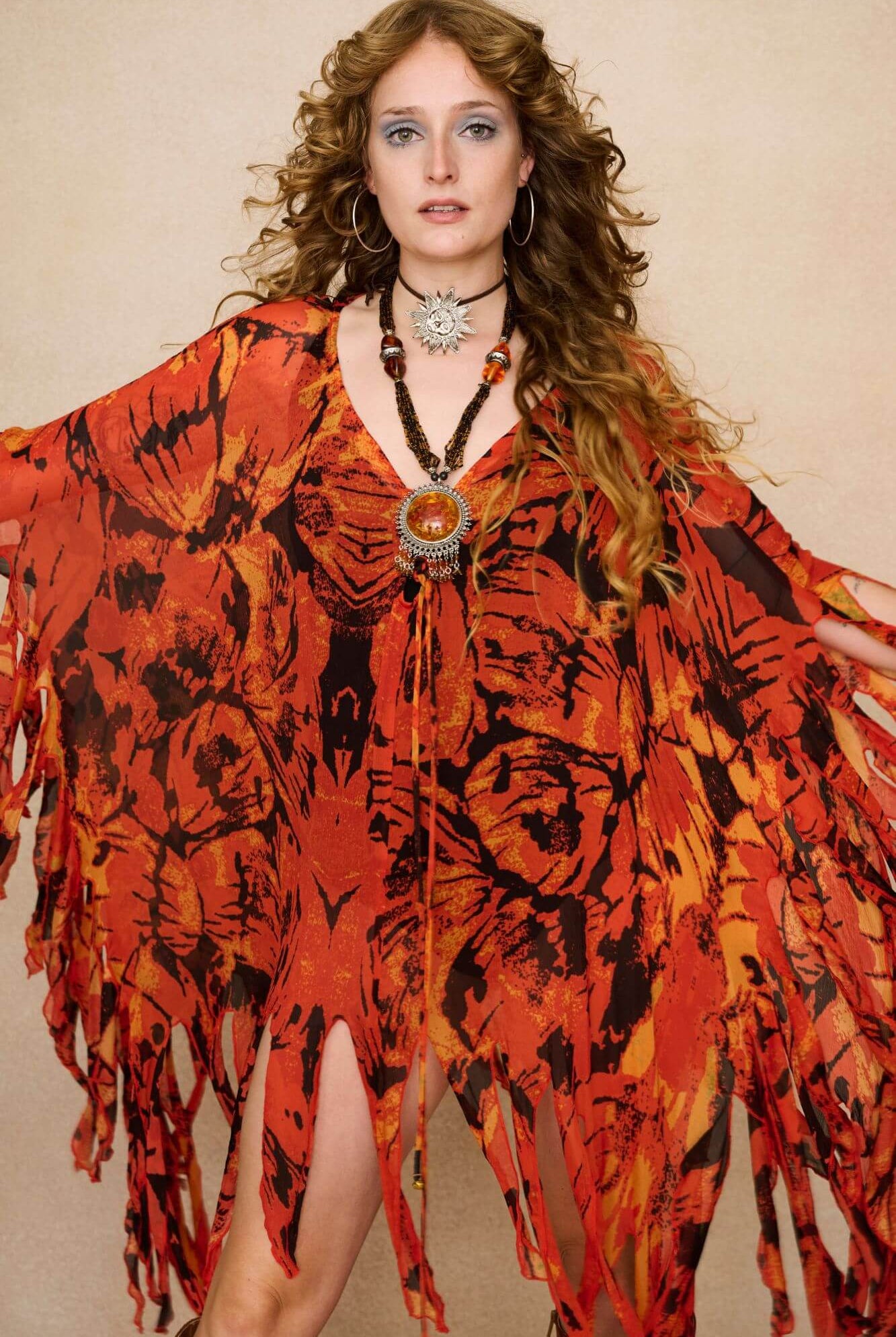Model wearing a vibrant red and black patterned dress cape with fringe against a beige background - Close up view