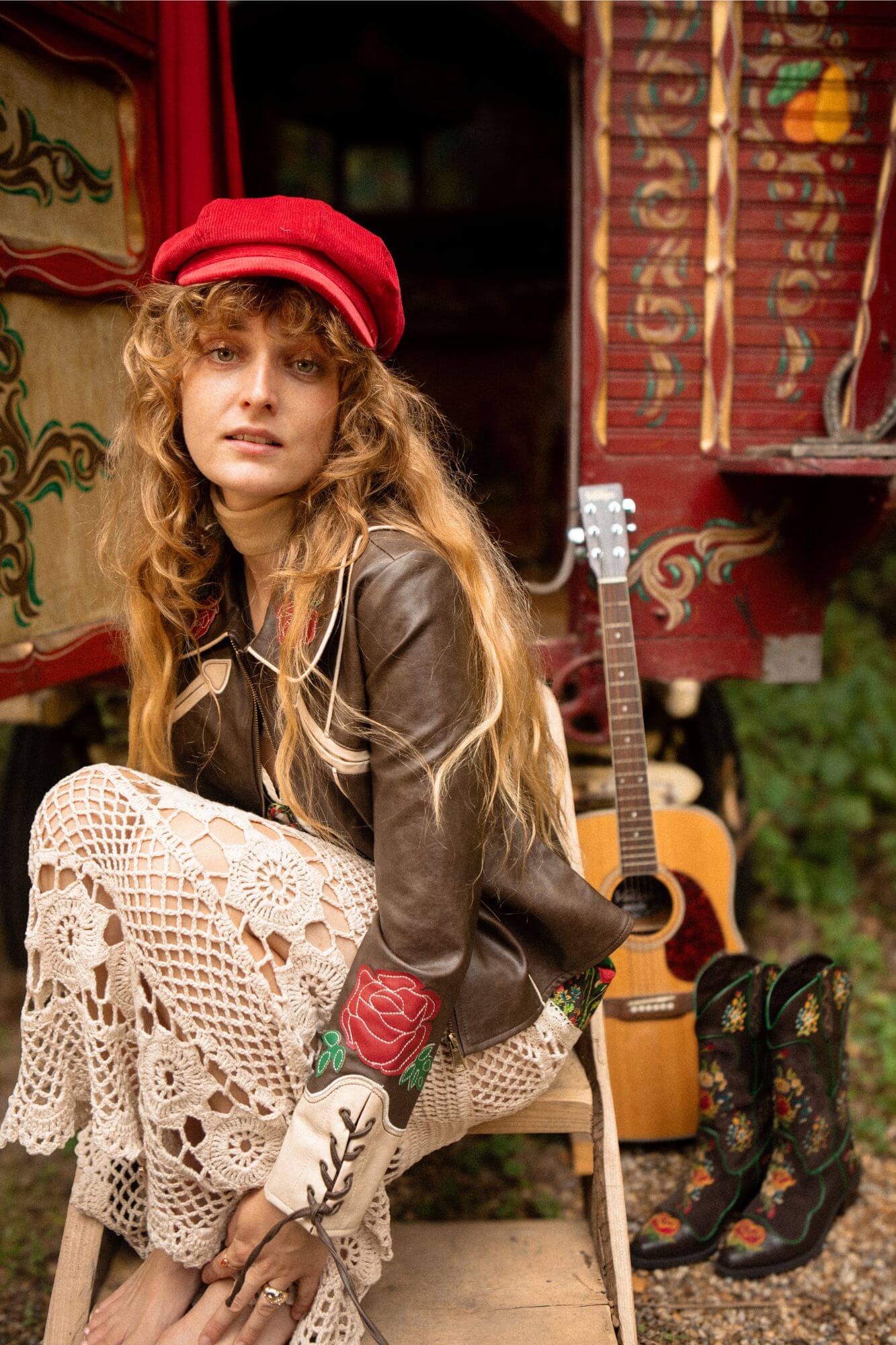 Woman in a red cap and leather jacket sitting with a guitar and boots in front of a colorful backdrop