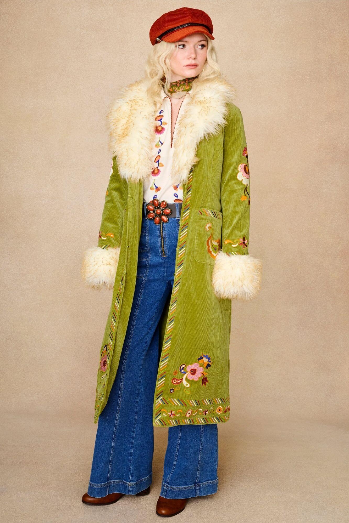 Model wears green embroidered Penny Lane coat in cotton corduroy with faux fur trim. A 70s-inspired statement coat made for bohemian, vintage styling by The Hippie Shake - open side view with denim