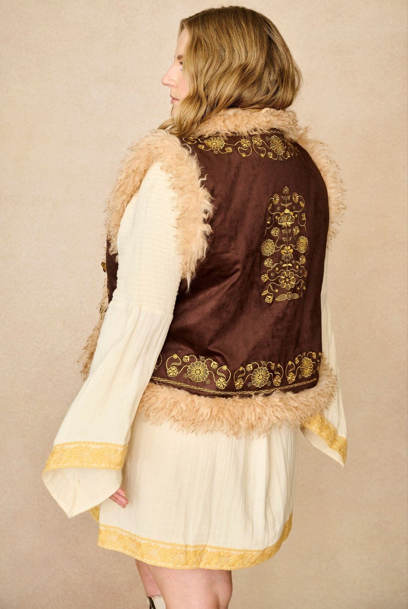 Model wears brown embroidered Penny Lane vest in faux suede with faux fur trim. A 70s-inspired boho layering piece made for vintage and festival styling by The Hippie Shake - back view paired with cream dress