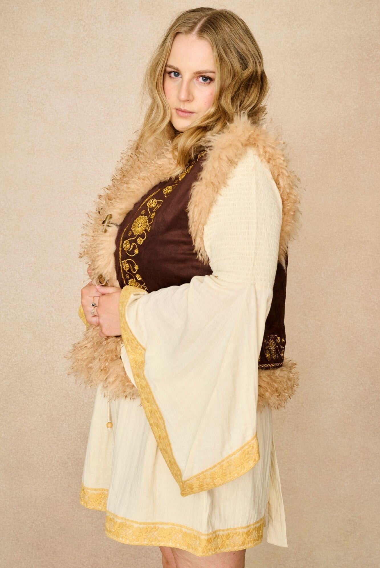 Model wears brown embroidered Penny Lane vest in faux suede with faux fur trim. A 70s-inspired boho layering piece made for vintage and festival styling by The Hippie Shake - side view paired with cream dress