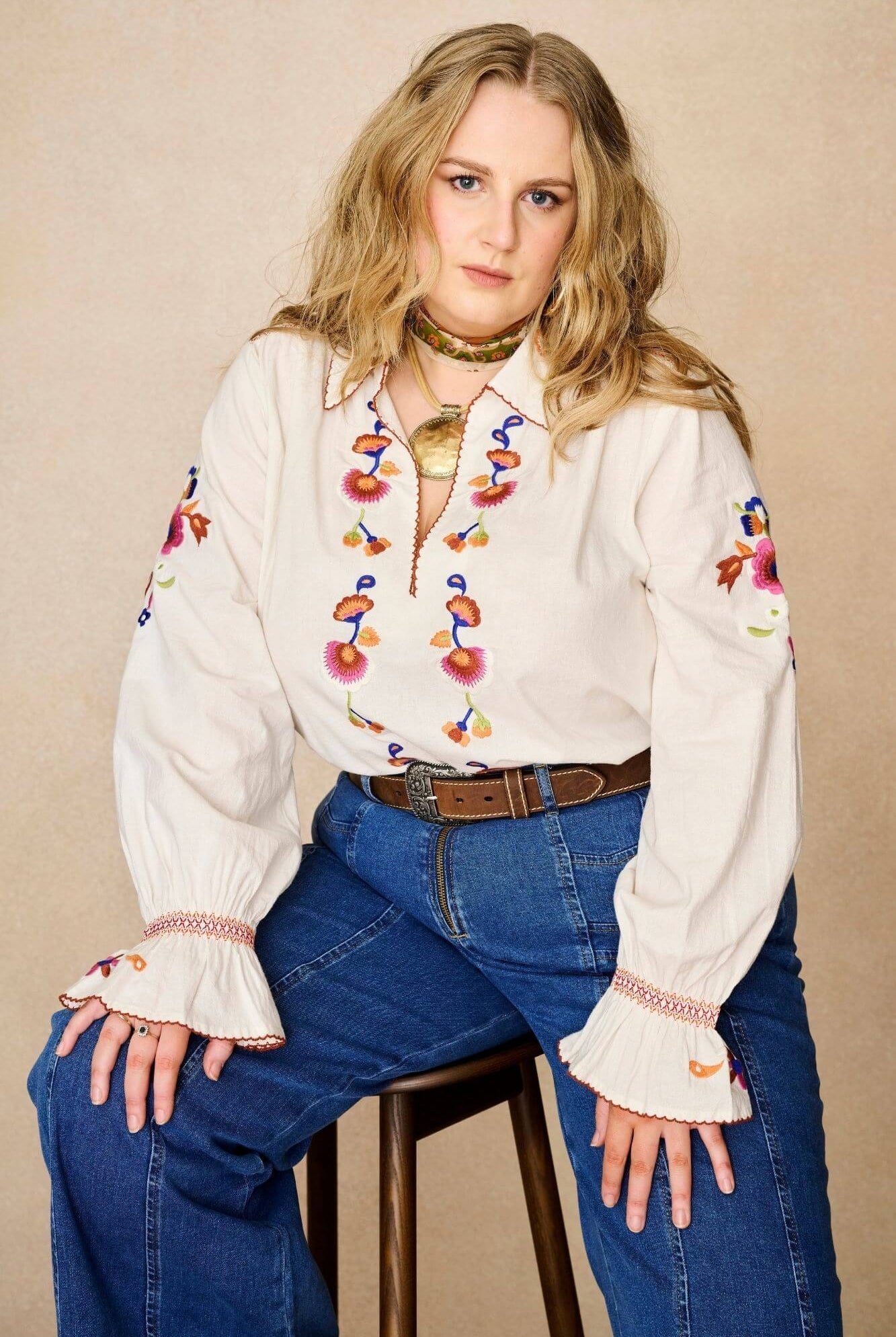 Model wears cream embroidered blouse in textured cotton with floral detailing and balloon sleeves paired with flared denim jeans. A 70s-inspired boho blouse made for everyday vintage styling by The Hippie Shake - sitting front view