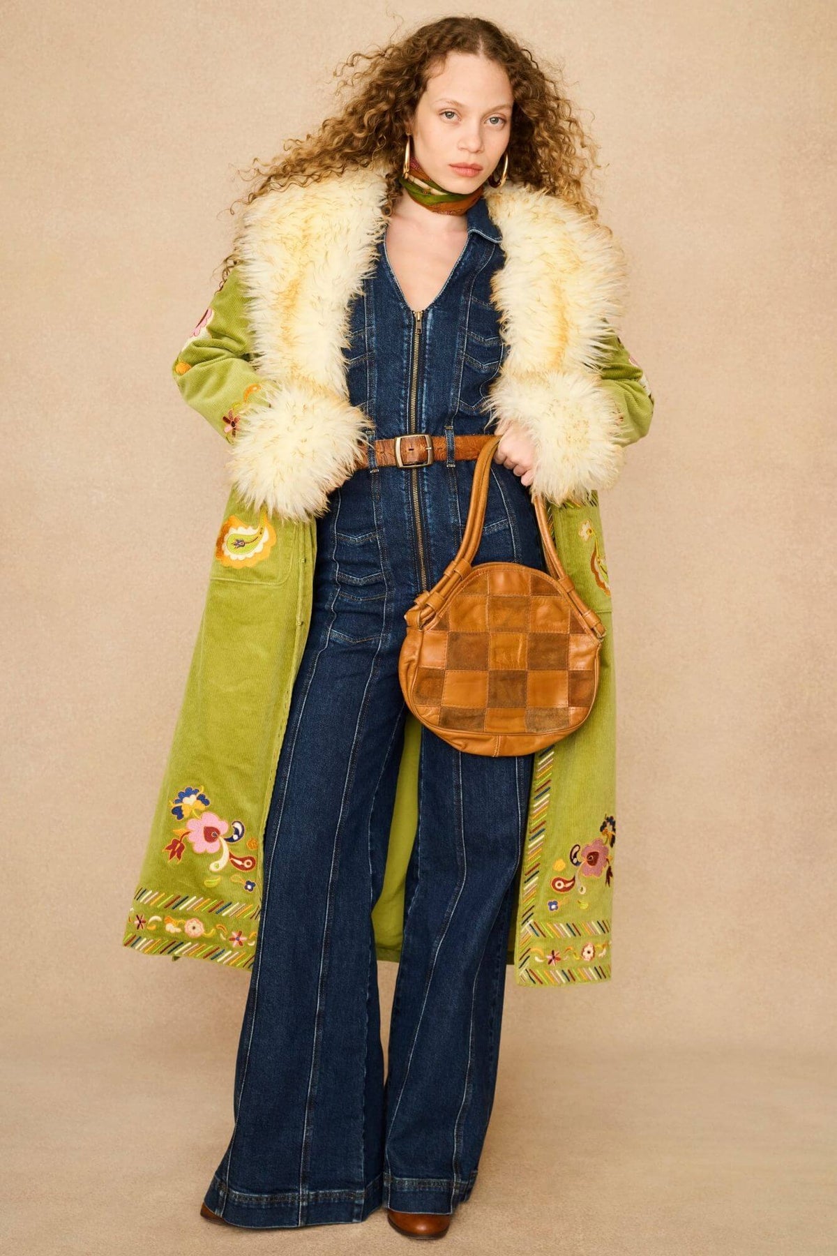 A model wearing a long olive green coat with a cream faux fur collar, a dark blue denim jumpsuit, and a brown woven round handbag, front view.