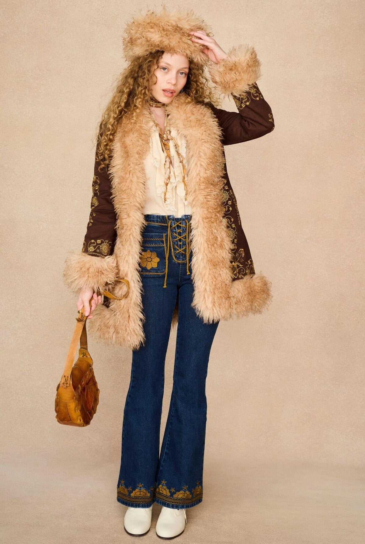 A model wearing a 70s bohemian-style outfit featuring a brown penny lane jacket with beige fur trim and a matching fur-trimmed hat.