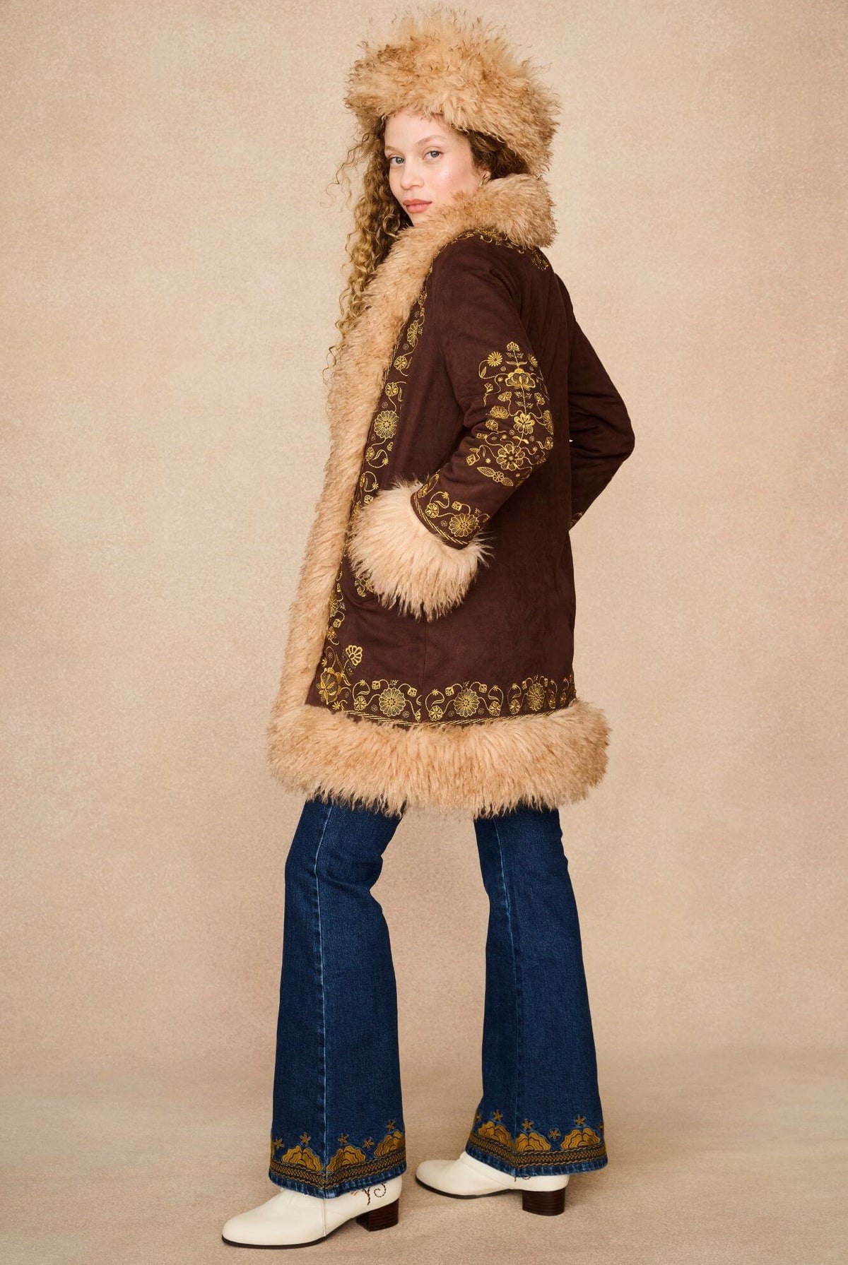 A model wearing a 70s bohemian-style outfit featuring a brown penny lane jacket with beige fur trim and a matching fur-trimmed hat.