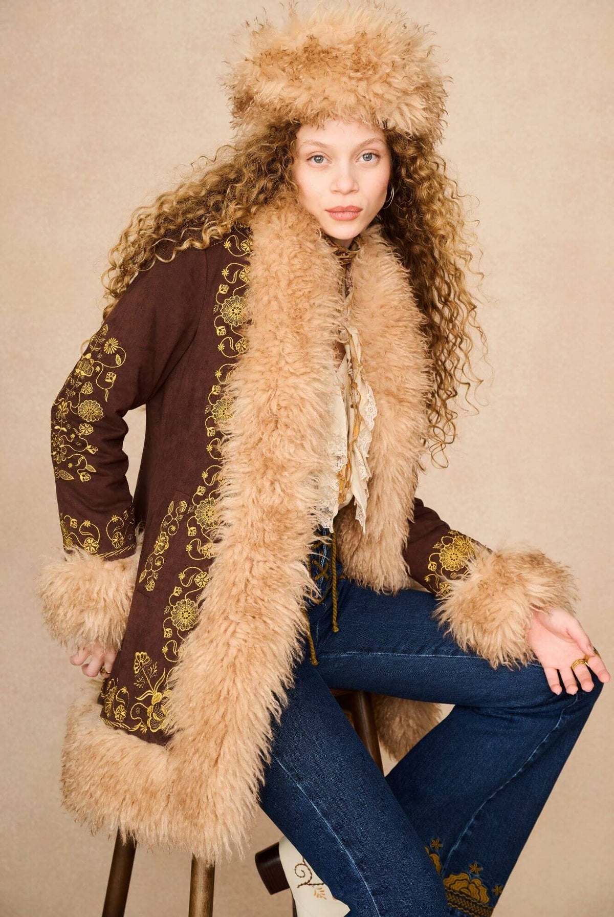 A model wearing a 70s bohemian-style outfit featuring a brown penny lane jacket with beige fur trim and a matching fur-trimmed hat.