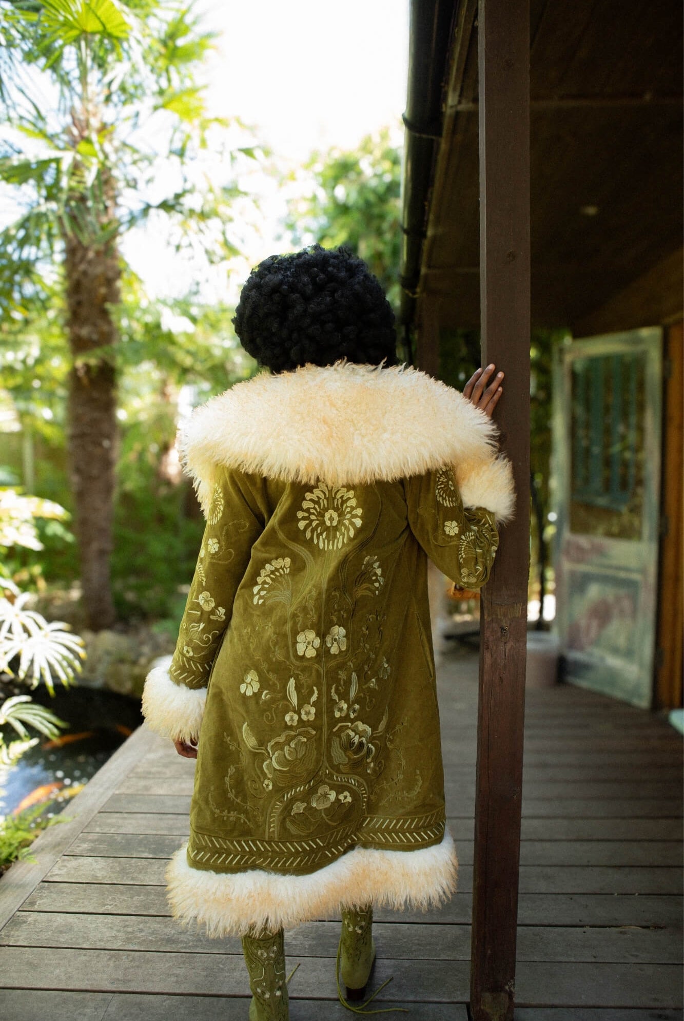Model viewed from behind wearing a long olive green Penny Lane embroidered coat with a large cream fur collar and hem, standing on a wooden deck outdoors near palm trees and a porch.