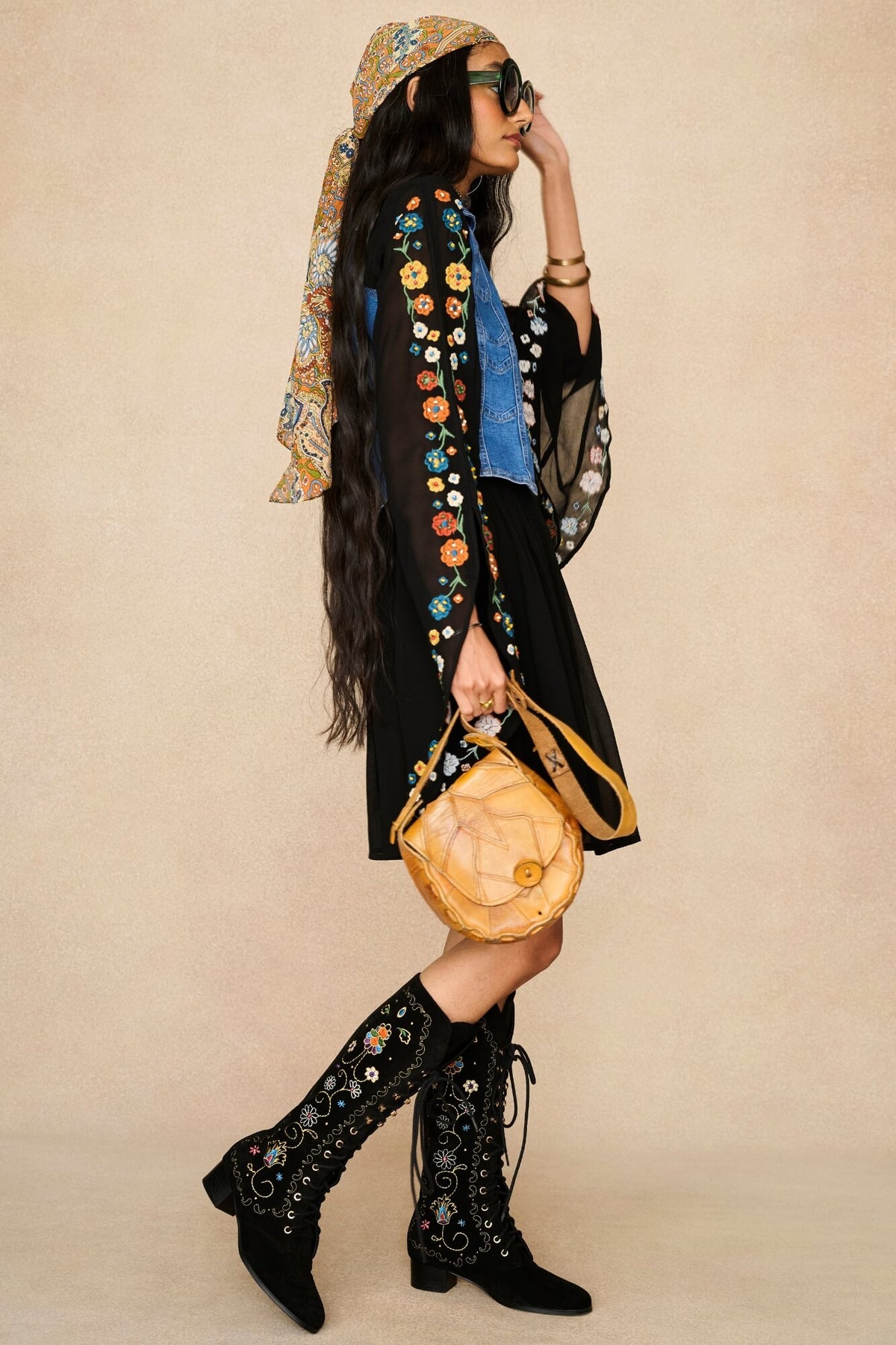 A woman models a black embroidered bohemian-style dress with a blue shirt underneath, a patterned headscarf, black lace-up knee-high boots, and a round tan leather shoulder bag, set against a solid beige studio background.