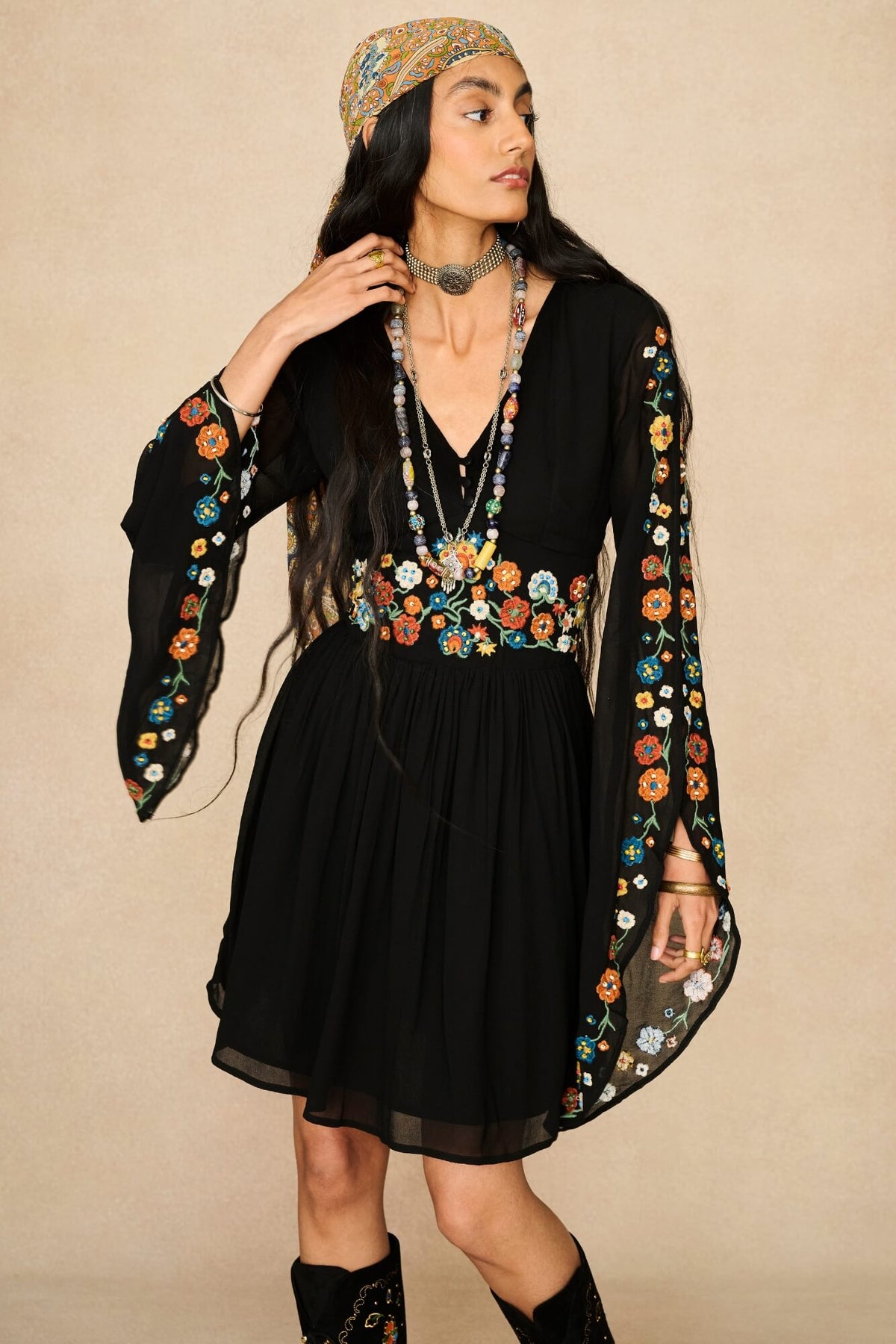 A woman modeling a black bohemian-style mini dress by The Hippie Shake with colourful floral embroidery on the sleeves and waist, paired with a matching patterned headscarf and layered necklaces, set against a plain beige background.