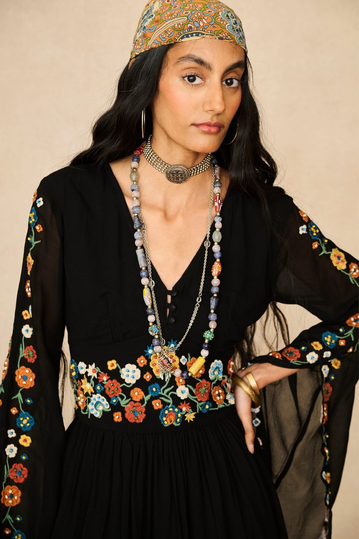 A woman wearing a black embroidered dress with colorful floral patterns on the sleeves and waist, accessorized with layered necklaces, a headpiece, and gold jewelry, set against a plain beige background.