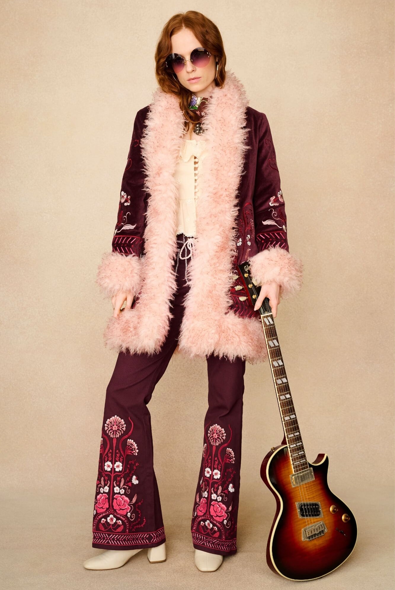Person in a vintage-style outfit with floral patterns, holding a guitar against a beige background