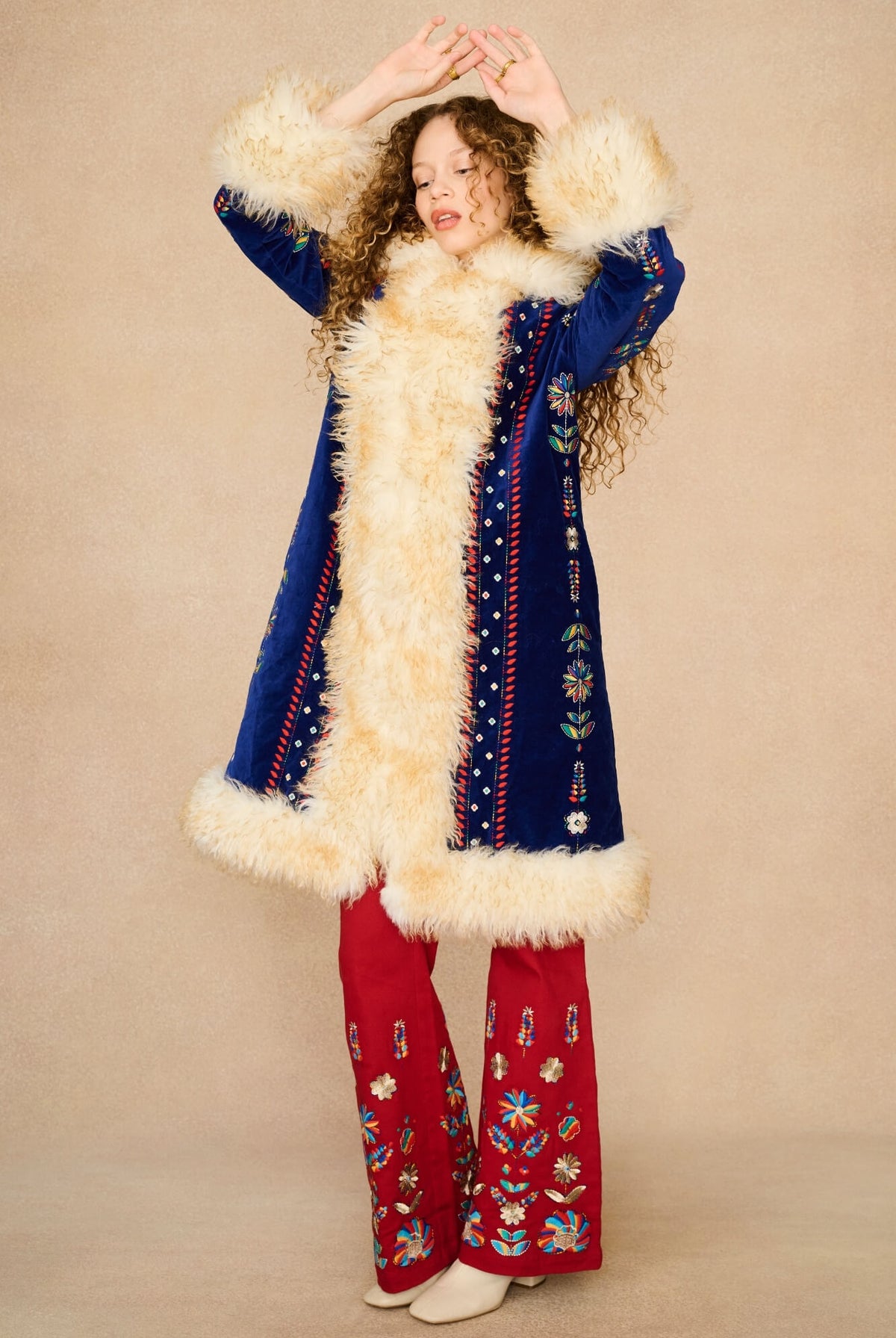 Model in a vintage-style bohemian Penny Lane Coat by The Hippie Shake with cream faux-fur trim, blue embroidered panels, and red embroidered pants against a solid beige background.