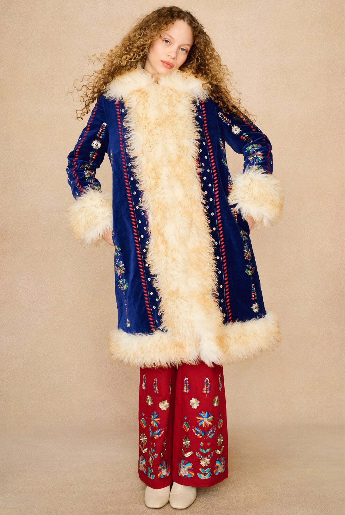 A model wearing a long blue velvet Penny Lane coat with a thick cream fur collar and cuffs, paired with red wide-leg pants featuring colorful embroidery, set against a plain beige studio background.